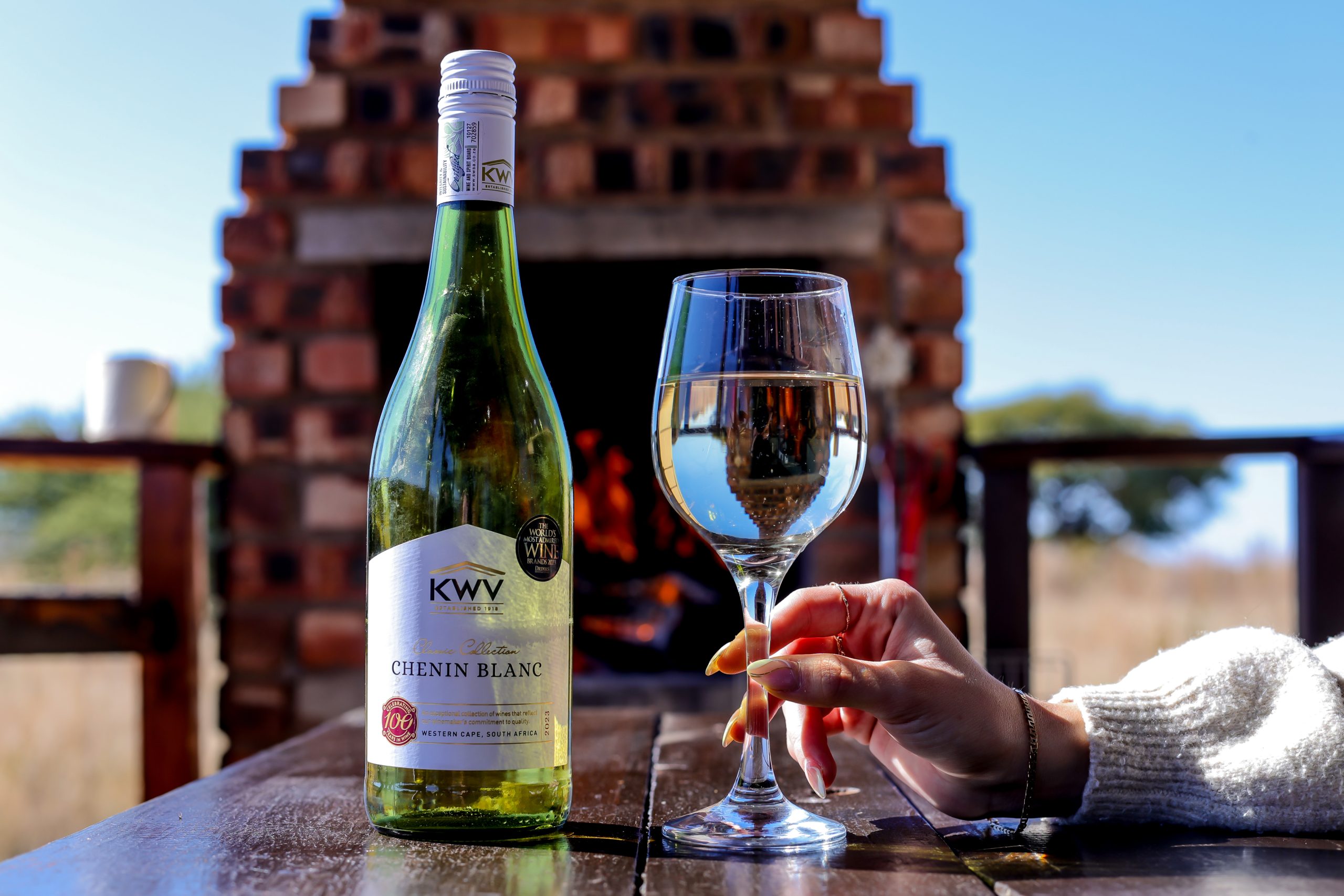 A close-up shot of a hand holding a glass of KWV Chenin Blanc, a bottle of the wine and a traditional South African braai area in the background, symbolising the relaxing and indulgent experience at the lodge.