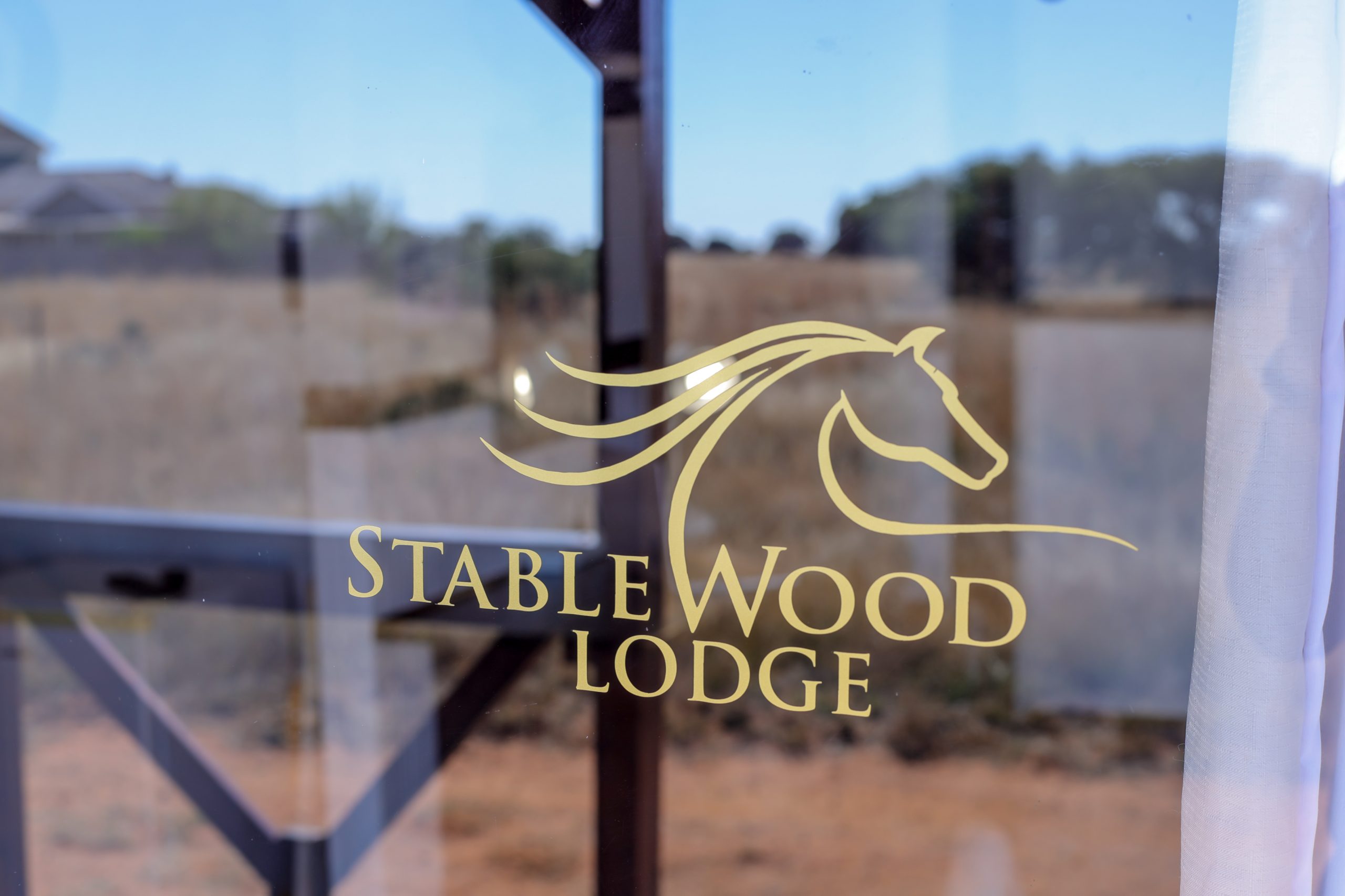 A close-up shot of the Stablewood Lodge logo, a stylised running horse with its name below it, printed on a clear glass door, with the blurred countryside visible in the background.