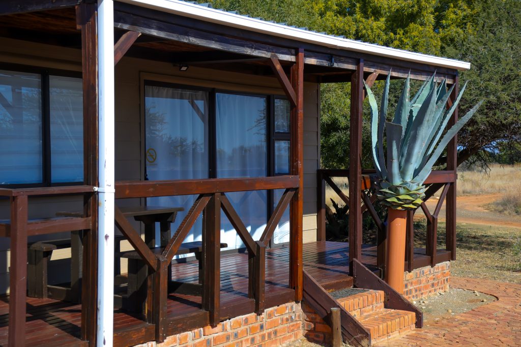 A self-catering chalet at Stablewood Lodge featuring a private wooden veranda with seating and a distinctive agave plant in a pot, set on a brick base, with glimpses of the surrounding Northern Cape vegetation.